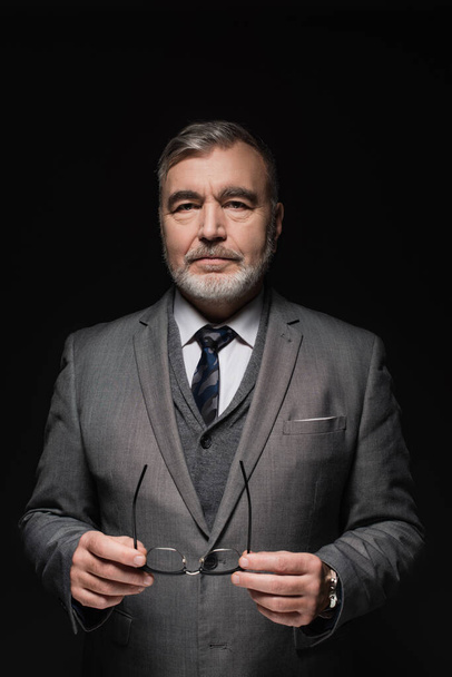 senior bearded businessman in grey suit holding eyeglasses and looking at camera isolated on black - Fotó, kép