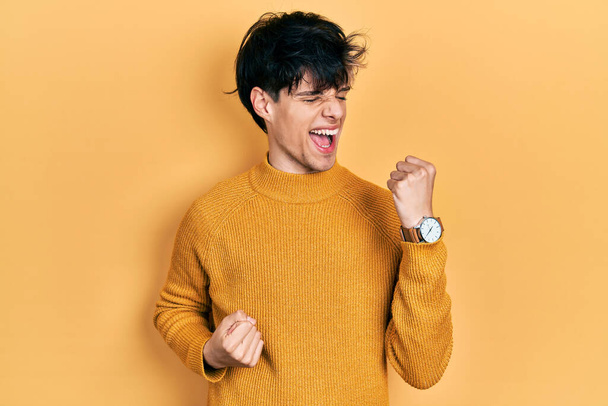 Bel giovane hipster con indosso maglione giallo casual che celebra sorpreso e stupito per il successo con le braccia alzate e gli occhi chiusi. concetto di vincitore.  - Foto, immagini
