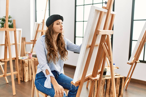 Mladý hispánský umělec žena malba na plátně v ateliéru umění hledá na stranu, relaxační profil pózovat s přirozenou tváří a sebevědomý úsměv.  - Fotografie, Obrázek