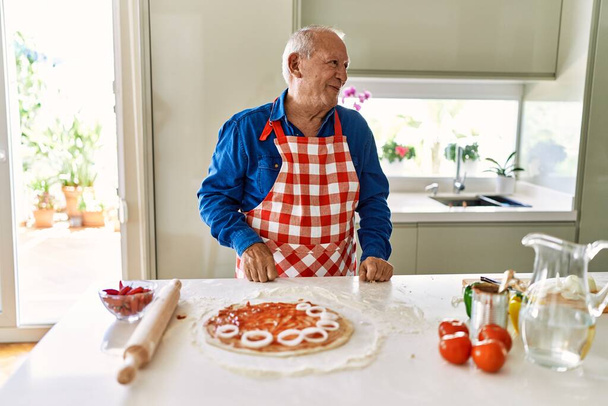 Senior man with grey hair cooking pizza at home kitchen looking away to side with smile on face, natural expression. laughing confident.  - Foto, afbeelding