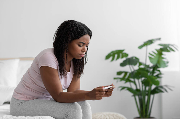 Despaired sad millennial african american lady sit on bed and holding pregnancy test in bedroom interior, profile - 写真・画像