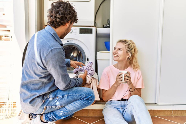 Young couple smiling happy drinking coffee while doing laundry at home. - Photo, Image