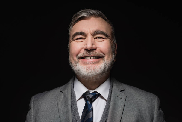 cheerful bearded man in formal wear smiling at camera isolated on black - Foto, afbeelding