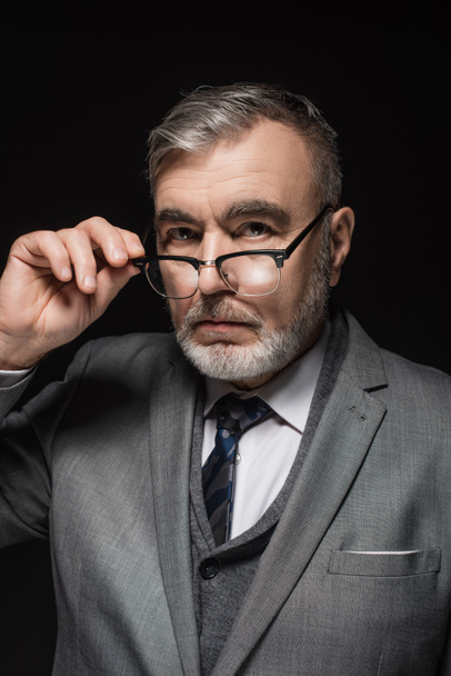 serious senior businessman adjusting eyeglasses while looking at camera isolated on black - Φωτογραφία, εικόνα