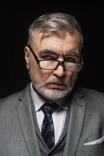 serious senior businessman in eyeglasses looking at camera isolated on black - Фото, изображение