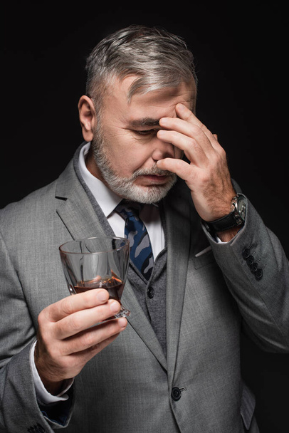 exhausted senior man holding glass of whiskey while suffering from headache isolated on black - Fotó, kép