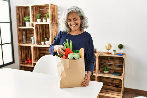 Mujer de pelo gris de mediana edad sosteniendo bolsa de papel con comestibles de pie en casa. - Foto, imagen