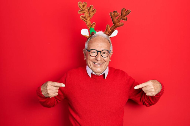 Старший чоловік з сірим волоссям, одягнений в різдвяний капелюх оленя, впевнено виглядає з посмішкою на обличчі, вказуючи на себе пальцями гордим і щасливим
.  - Фото, зображення
