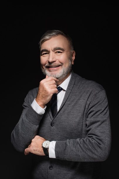 happy senior man in jumper and tie smiling at camera isolated on black - Foto, immagini