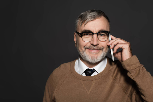 smiling man in beige jumper and eyeglasses talking on cellphone isolated on grey - Fotografie, Obrázek