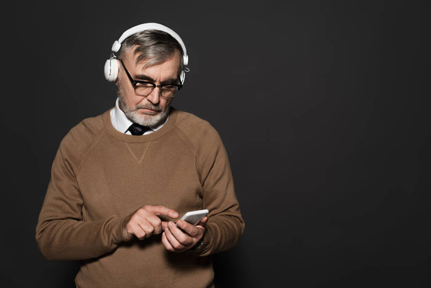 senior man in beige jumper and headphones pointing at mobile phone on dark grey - Fotoğraf, Görsel