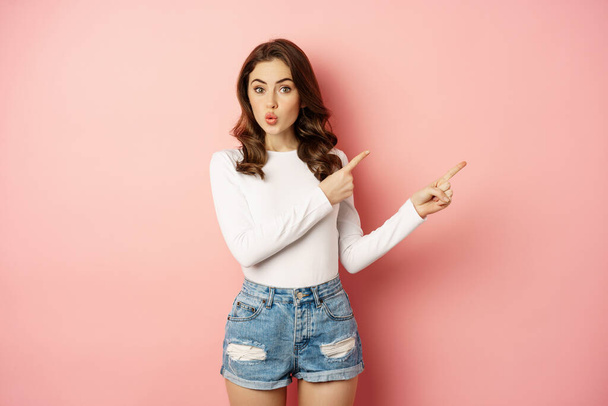 Portrait of intrigued stylish girl showing interest in advertisement, look curious, pointing at upper right corner logo, banner or discount news, pink background - Foto, Imagem
