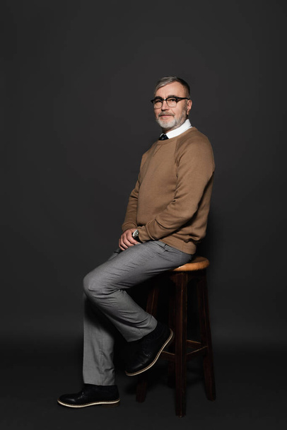 full length view of senior man in stylish clothes sitting on high stool on dark grey - Photo, image