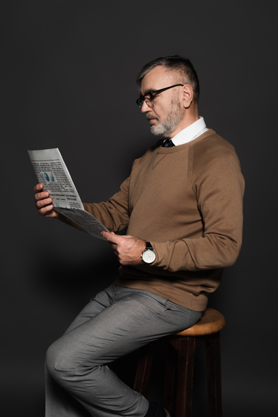 stylish senior man in eyeglasses reading newspaper while sitting on stool on dark grey - Photo, Image