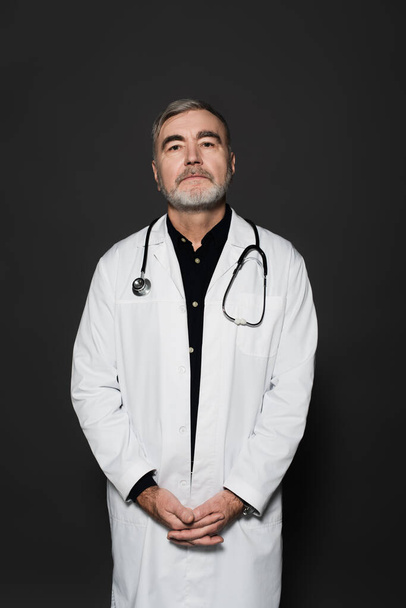 bearded doctor in white coat standing with clasped hands and looking at camera isolated on dark grey - Fotoğraf, Görsel