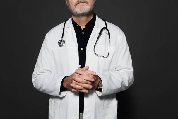 cropped view of senior doctor with stethoscope on neck standing with clasped hands isolated on dark grey - Fotoğraf, Görsel