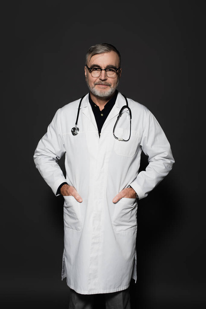 smiling senior doctor looking at camera while standing with hands in pockets on dark grey - Foto, afbeelding