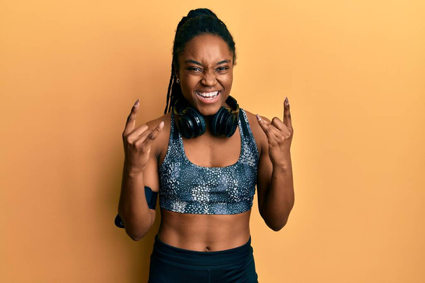 African american woman with braided hair wearing sportswear and arm band shouting with crazy expression doing rock symbol with hands up. music star. heavy concept.  - Photo, Image