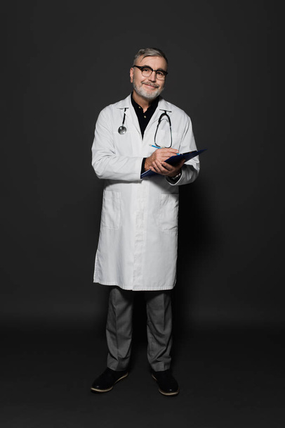 full length view of happy senior doctor in white coat writing on clipboard on dark grey - Foto, Imagen