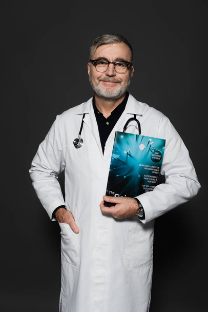 mosolygós orvos szemüveg áll magazin és kéz a zsebben fehér kabátot elszigetelt sötétszürke - Fotó, kép