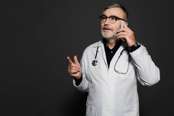 bearded doctor looking away and pointing with finger while talking on cellphone on dark grey - Foto, Imagem
