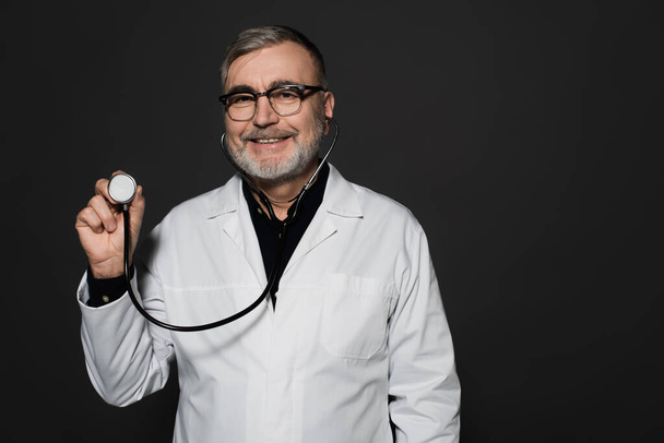 cheerful senior doctor in eyeglasses holding stethoscope isolated on black - Fotografie, Obrázek