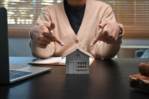 Молодая женщина указывает на модель дома на столе. Концепция инвестиций в ипотеку и недвижимость. - Фото, изображение