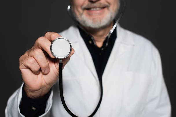 cropped view of blurred smiling physician holding stethoscope isolated on black - Foto, Imagen