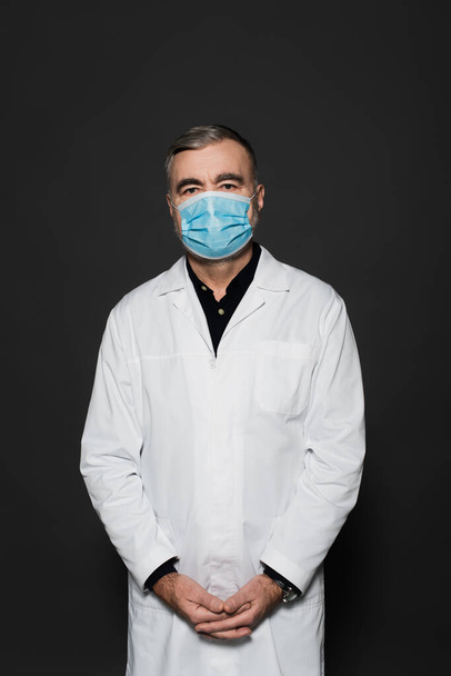 senior physician in white coat and medical mask looking at camera isolated on dark grey - 写真・画像
