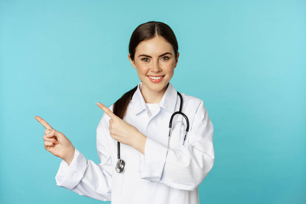 Portrait of smiling medical worker, girl doctor in white coat with stethoscope, pointing fingers left, showing medical clinic advertisement, torquoise background - Фото, изображение