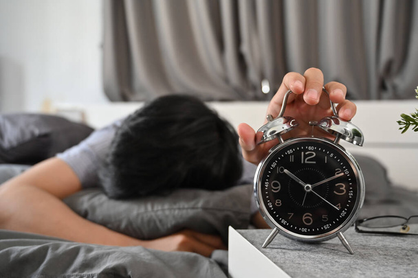 Молодой человек лежит в постели и выключает будильник. - Фото, изображение