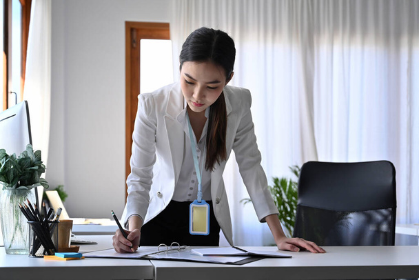 Affascinante donna d'affari in piedi alla scrivania del suo ufficio e controllare i rapporti. - Foto, immagini