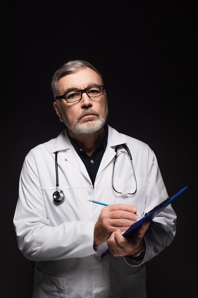 senior doctor in eyeglasses writing on clipboard while looking at camera isolated on black - Foto, Imagen