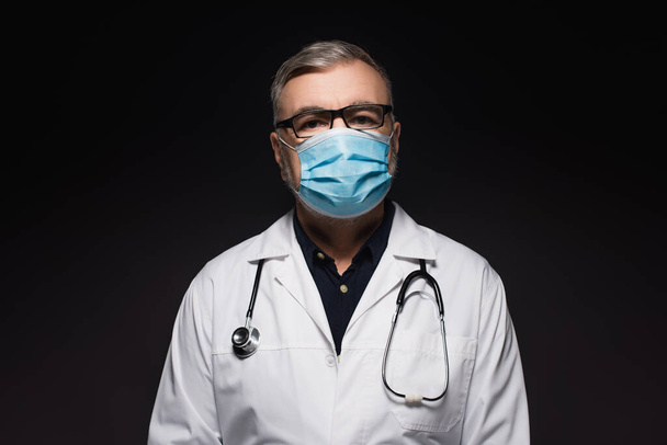 senior doctor in white coat and medical mask looking at camera isolated on black - Foto, afbeelding