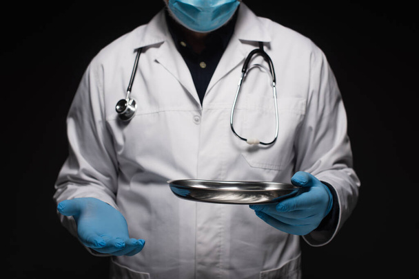 καλλιεργημένη άποψη του γιατρού με ιατρική μάσκα και γάντια λατέξ που κρατούν ανοξείδωτο ιατρικό δίσκο απομονώνονται σε μαύρο - Φωτογραφία, εικόνα