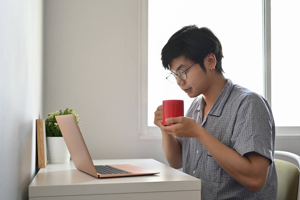 Peaceful asian man in pajamas using computer laptop in bedroom and drinking coffee. - Фото, зображення