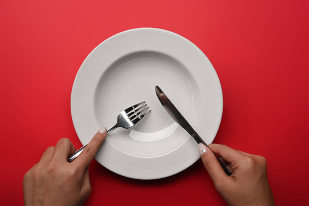 Woman holding fork and knife in hands with empty plate over red background. - Zdjęcie, obraz