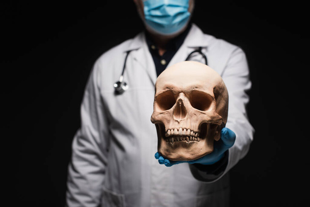 καλλιεργημένη άποψη θαμπής γιατρού με ιατρική μάσκα που κρατά το κρανίο απομονωμένο σε μαύρο - Φωτογραφία, εικόνα