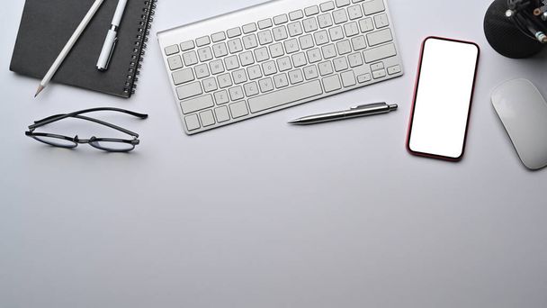 Espacio de trabajo moderno con teléfono inteligente, teclado inalámbrico y gafas sobre mesa blanca. - Foto, imagen