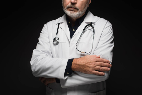 частковий вид бородатого лікаря зі стетоскопом на шиї, що стоїть з схрещеними руками, ізольовані на чорному
 - Фото, зображення