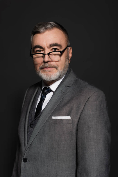 senior businessman in formal wear and eyeglasses looking at camera isolated on dark grey - Фото, изображение