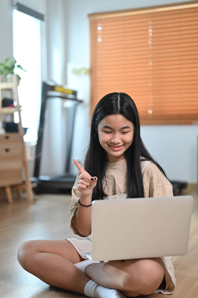 Усміхнена азіатська жінка має відеодзвінок з ноутбуком, сидячи на підлозі у вітальні
. - Фото, зображення