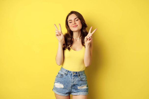 Positives Mädchen, weibliches Modell, das Frieden zeigt, V-Zeichen-Geste und Lächeln, in Tank-Top und Jeanshose stehend, gelber Hintergrund - Foto, Bild