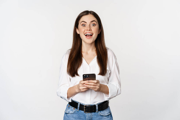 Young woman with smartphone, smiling and looking at camera, using mobile phone app, cellular technology and online shopping concept, white background - Φωτογραφία, εικόνα