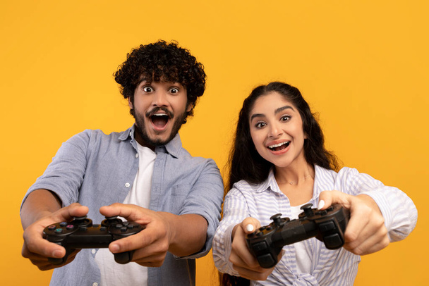 Concepto de estilo de vida. Feliz pareja india jugando videojuegos en línea, utilizando joystick gamepad, fondo amarillo - Foto, imagen