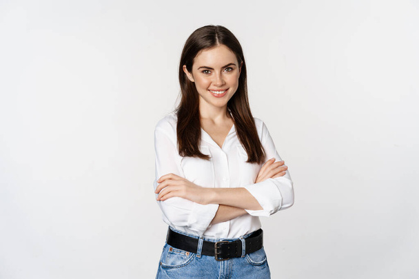 Smiling young businesswoman, female entrepreneur in white shirt, cross arms on chest like professional, standing over studio background - Fotó, kép