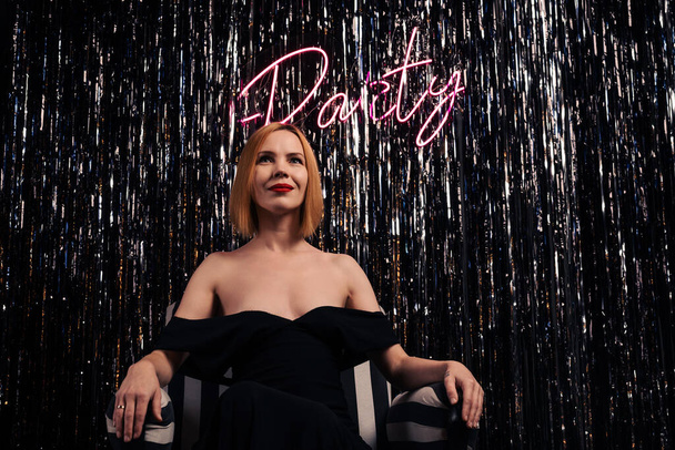 Stylish middle-aged woman sits in a chair. Party inscription on the luminous wall. Bare shoulders. A woman of forty years old. Clubbing, nightlife. Celebration, entertainment concept. Woman and party. - Foto, Imagen