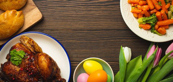 Comida de Pascua en mesa rústica oscura: huevos de colores pastel, pollo asado, verduras, bollos - Foto, imagen