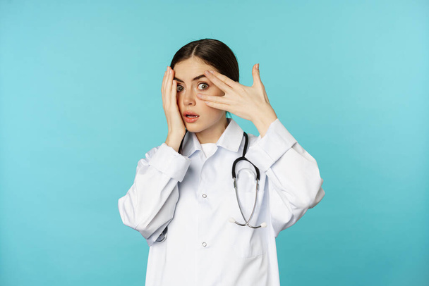 Потрясенная женщина-доктор, подглядывающая сквозь пальцы, отворачивающаяся от грязного уродства, стоящая в белом халате на торкизном фоне - Фото, изображение