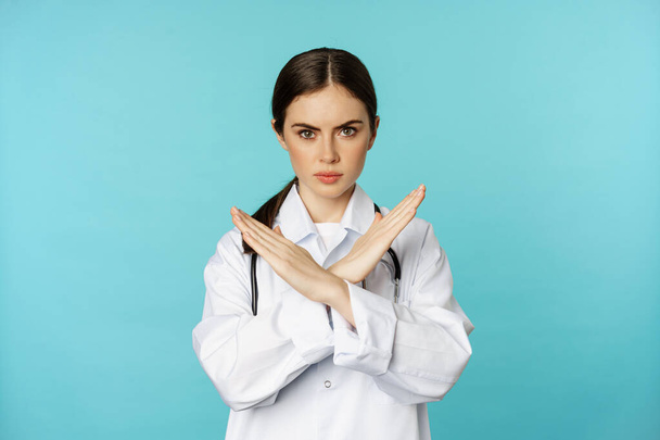 Ritratto di medico donna, medico mostrando croce, smettere di proibire gesto, vietare smth, in piedi in cappotto bianco su sfondo torchese - Foto, immagini